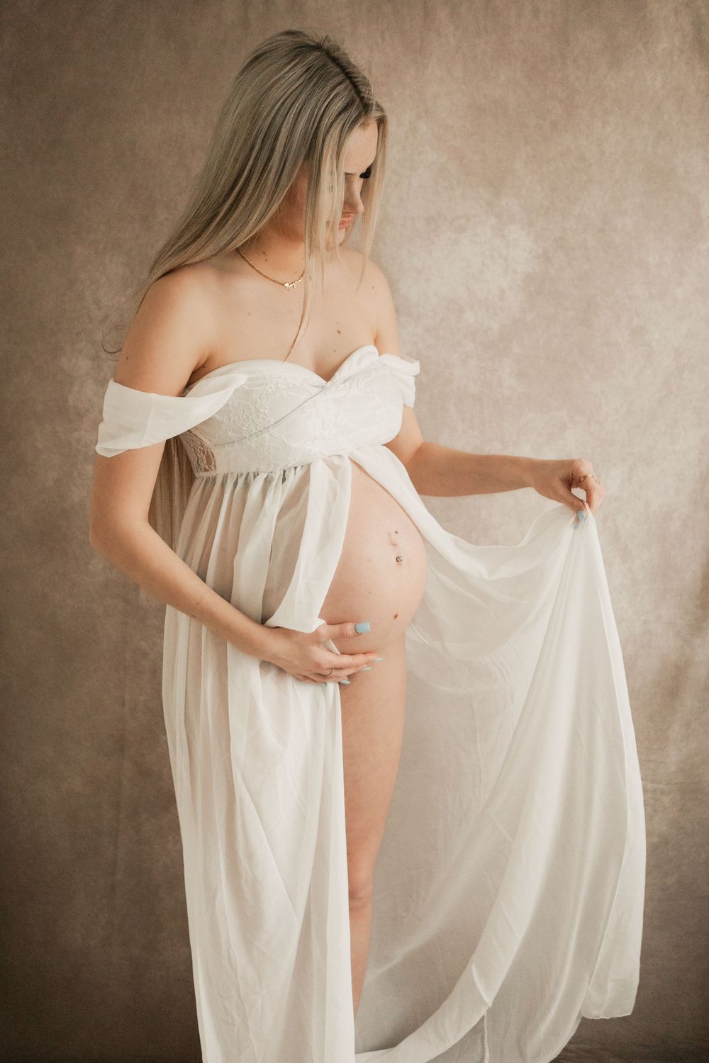 Pregnant woman in white off-shoulder sheer gown , showcasing baby bump in elegant maternity studio portrait taken by Cornwall Photographer