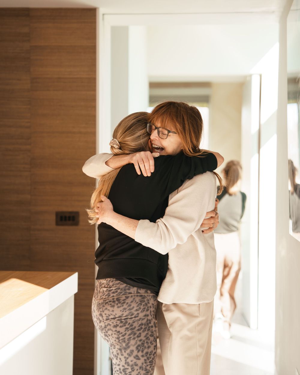Two women sharing a warm hug in a modern home interior, one wearing black top and leopard print pants.