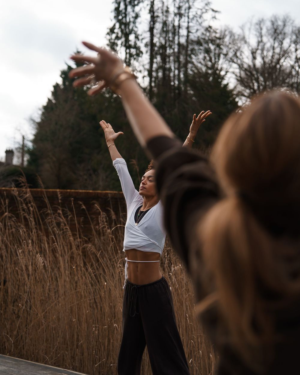 Two women dancing outdoors with arms raised near tall dry grass and trees in a natural setting.