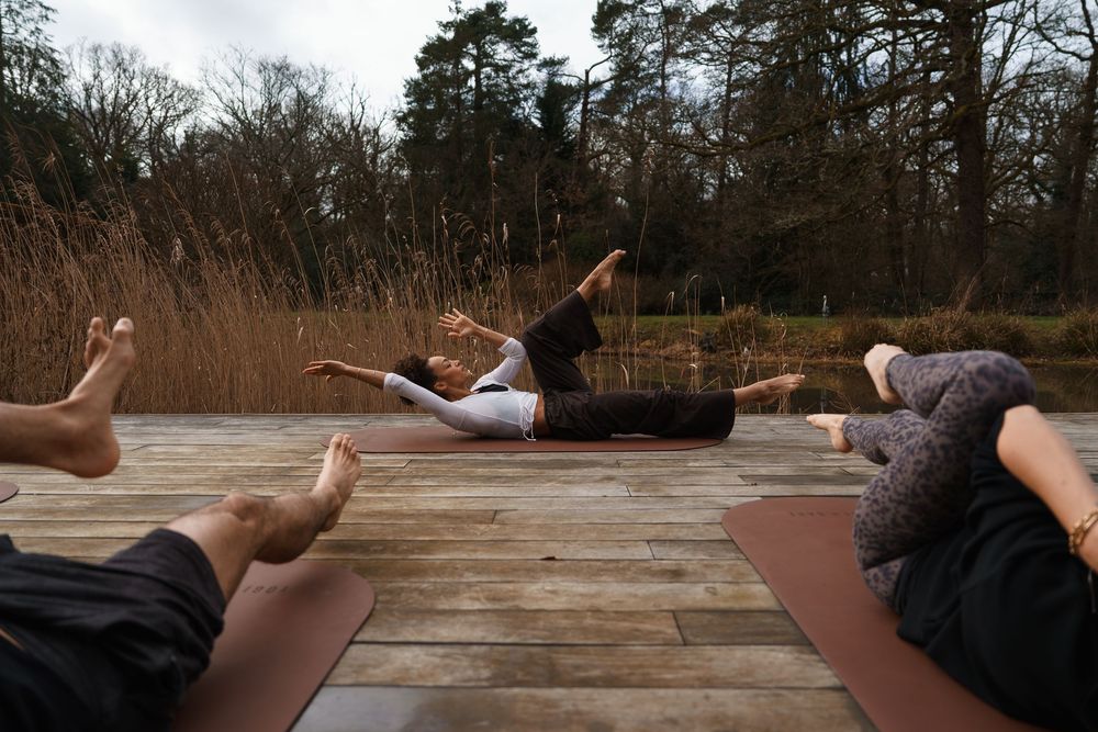 Group of people practicing yoga poses on outdoor wooden deck surrounded by autumn trees and tall grass.