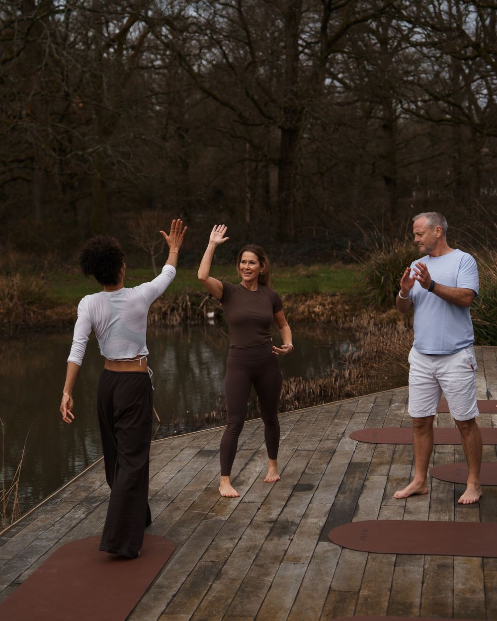 Three people practicing yoga on a wooden deck outdoors near a pond, surrounded by trees.