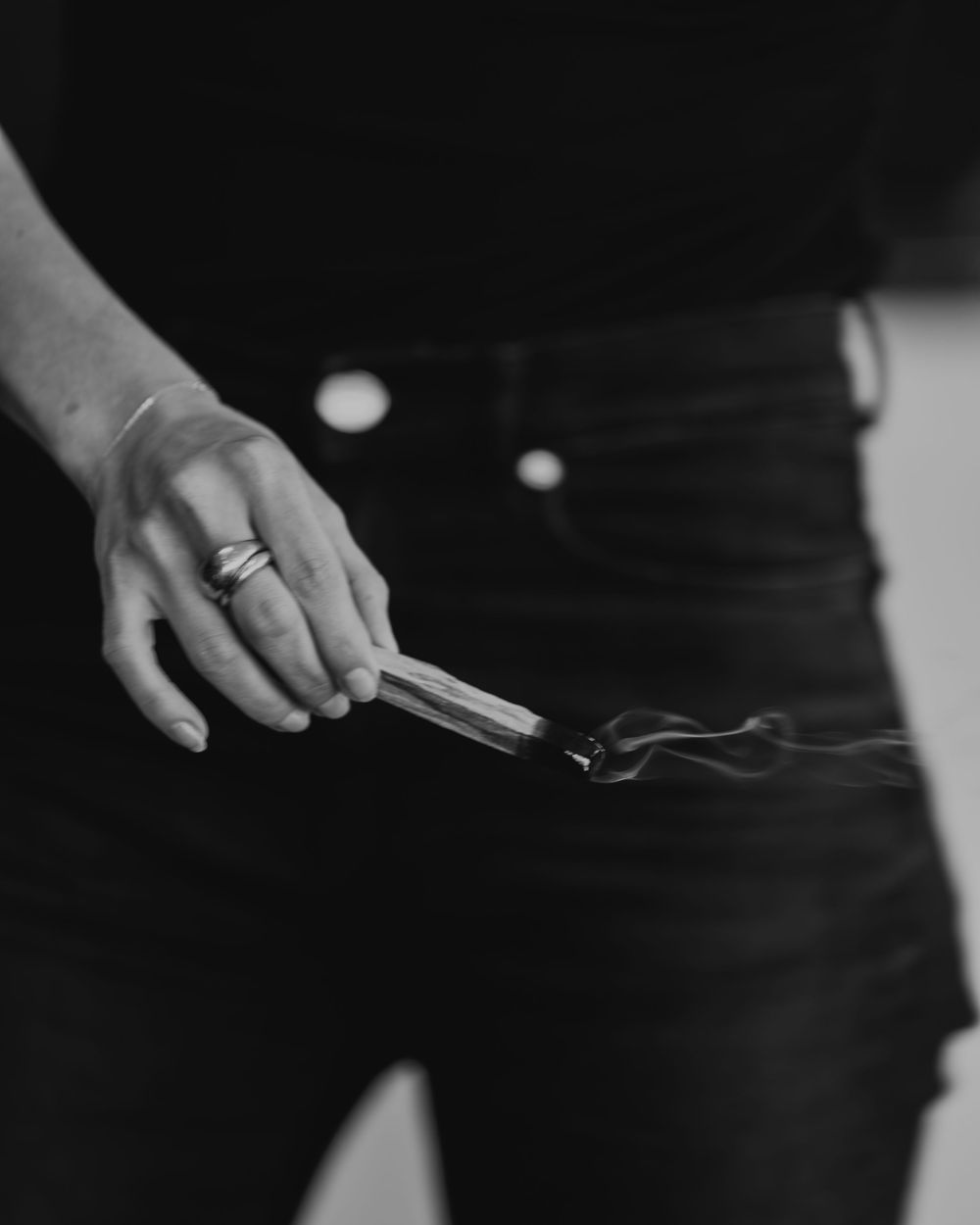 Black and white photo of a hand holding a smoking incense stick, wearing rings, dressed in black jeans.