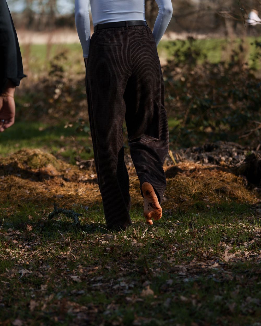 Person walking barefoot on grass wearing dark trousers and white top in an outdoor natural setting.