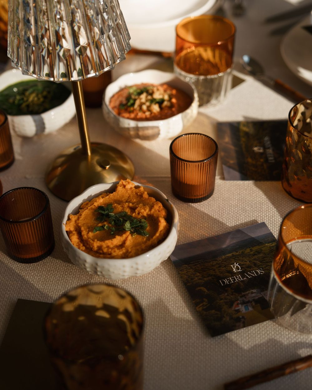 Elegant table setting with amber glassware, white bowls of dips, and a gold lamp on a linen tablecloth.