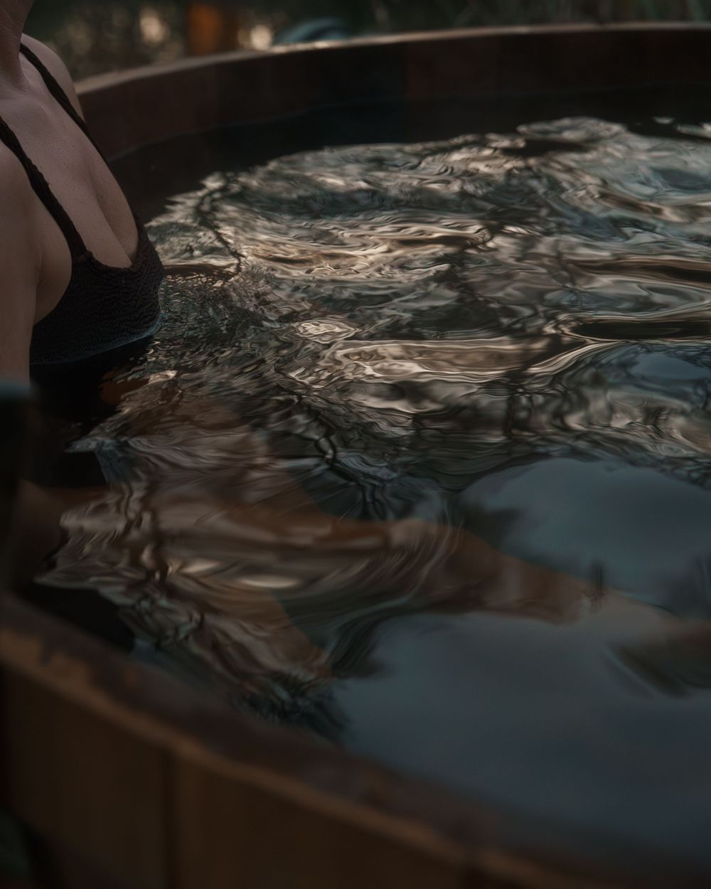 A person in a black swimsuit sits in a wooden barrel hot tub with dark reflective water.