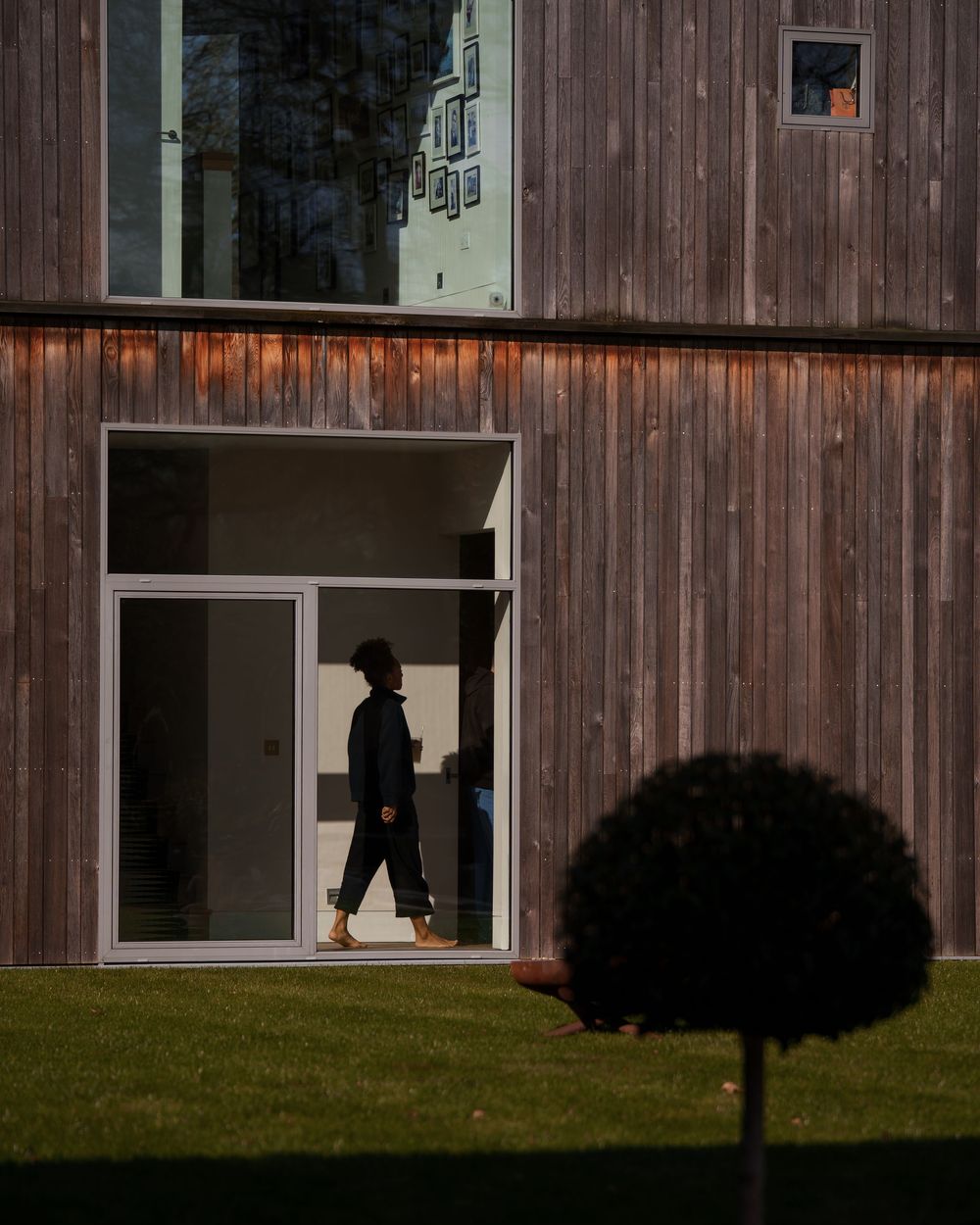 Person in dark clothing walking past a modern wooden clad building with large white framed windows on green lawn.