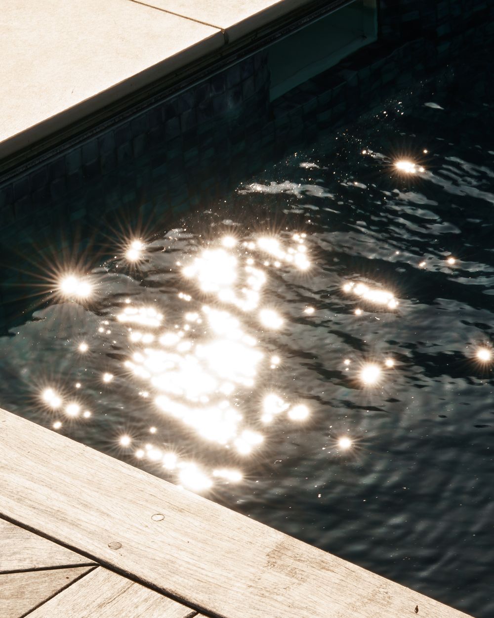 Sunlight reflects and sparkles on the dark water surface of an outdoor swimming pool with wooden decking.