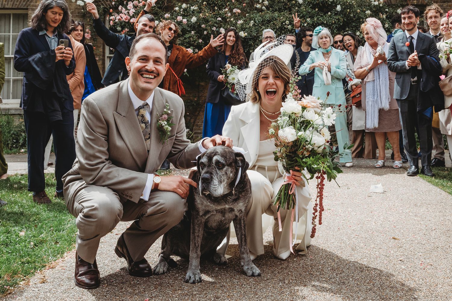 Bride in white pantsuit holding bouquet stands with groom in gray suit outdoors in lush green park setting.