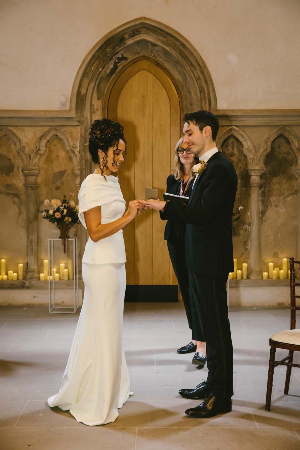 Bride placing wedding ring on groom's finger during elegant chapel ceremony with candles and gothic arched door.
