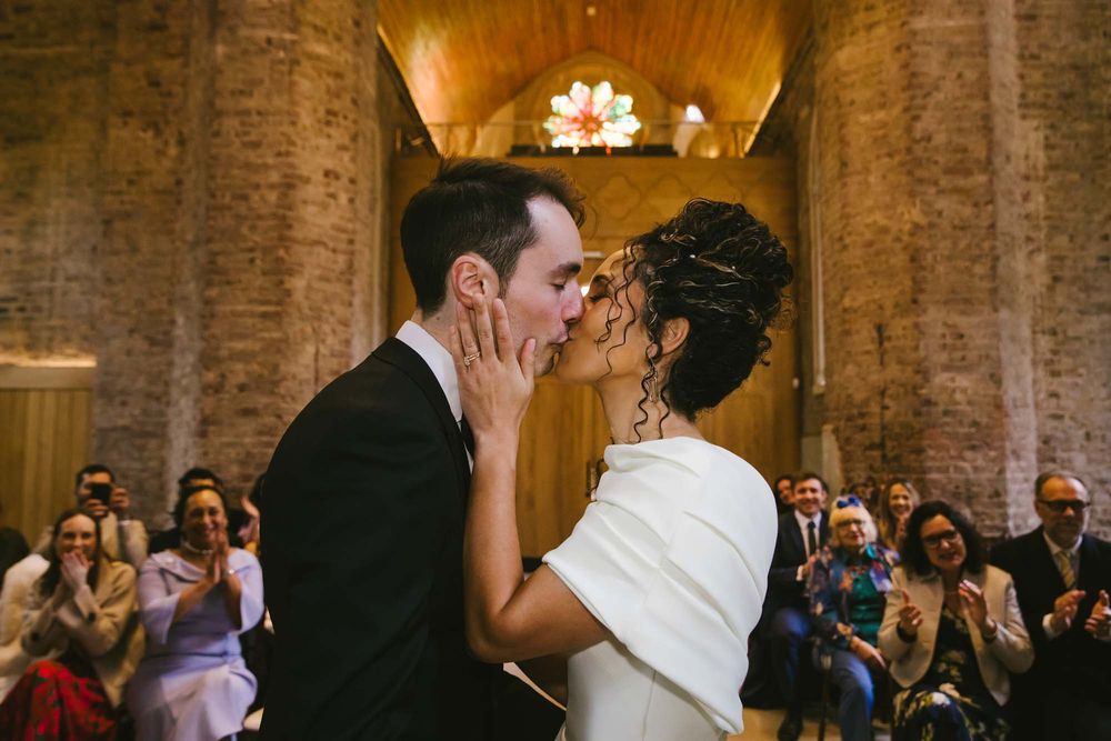 Bride and groom share first kiss at wedding ceremony while guests applaud in a rustic brick venue.