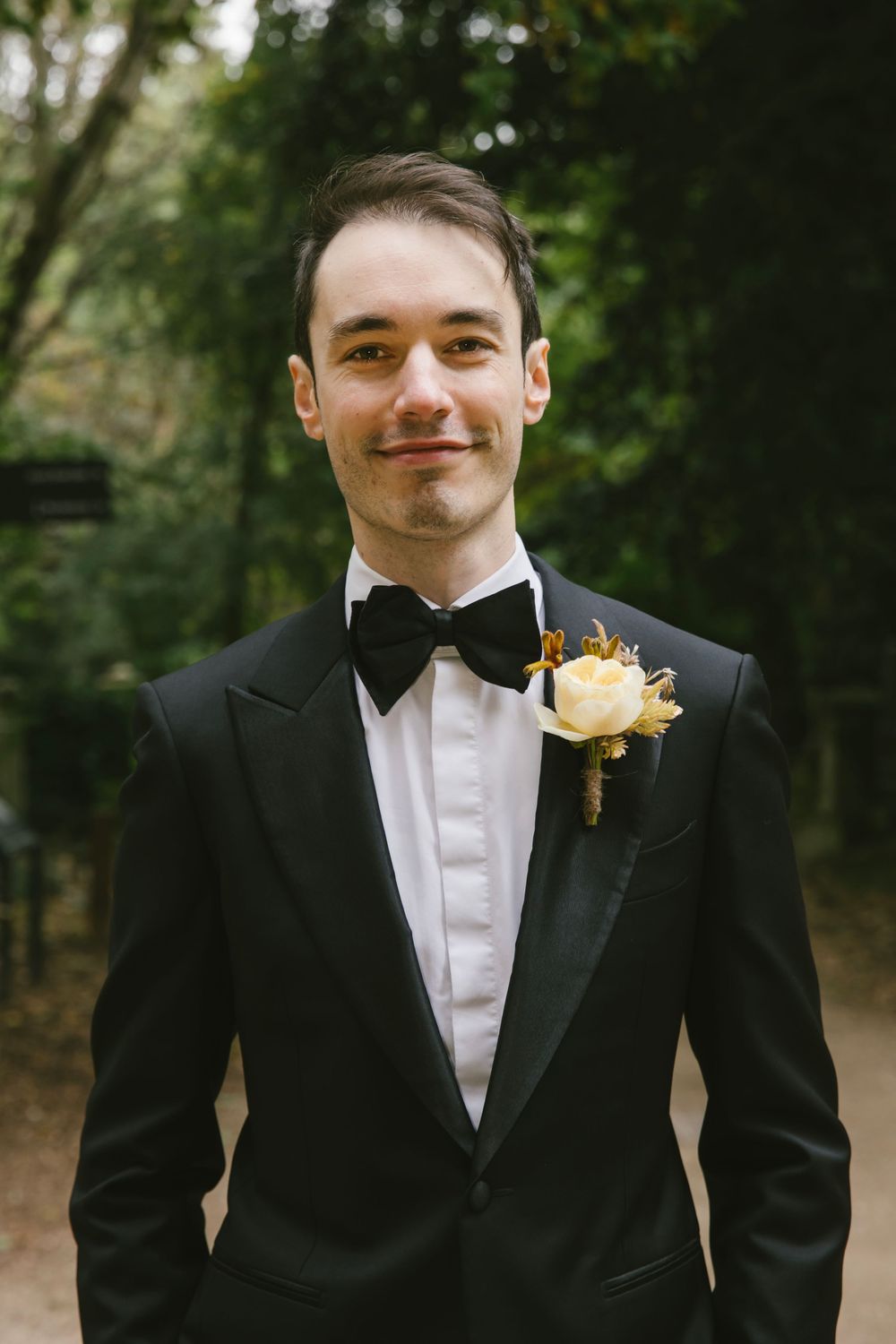 Groom in black tuxedo with bow tie and white rose boutonniere smiling outdoors among green trees.