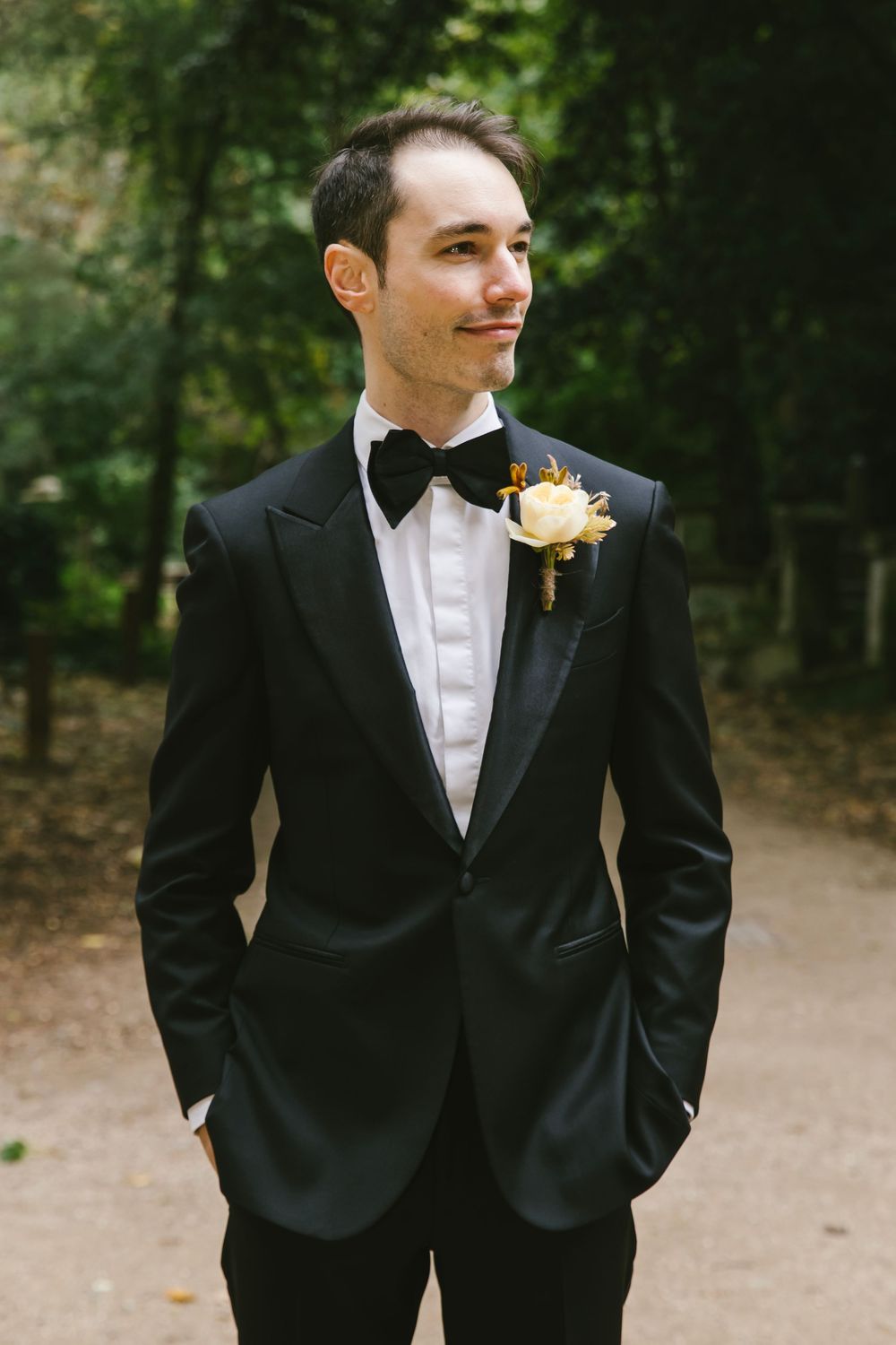 Groom in black tuxedo with bow tie and white boutonniere stands outdoors in a wooded setting.