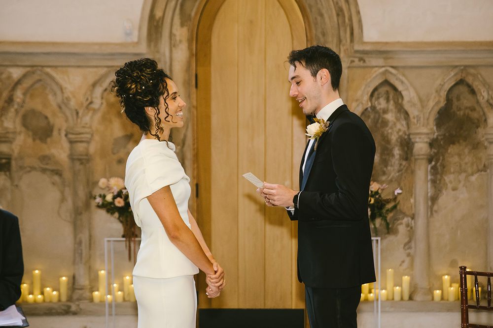 Groom reads wedding vows to bride at elegant chapel ceremony with candles and gothic wooden door backdrop.