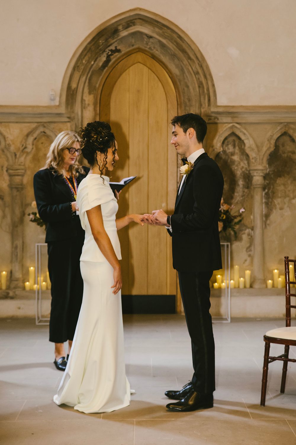 Couple exchanging vows at candlelit wedding ceremony in gothic chapel, officiant holding book, bride in white gown.