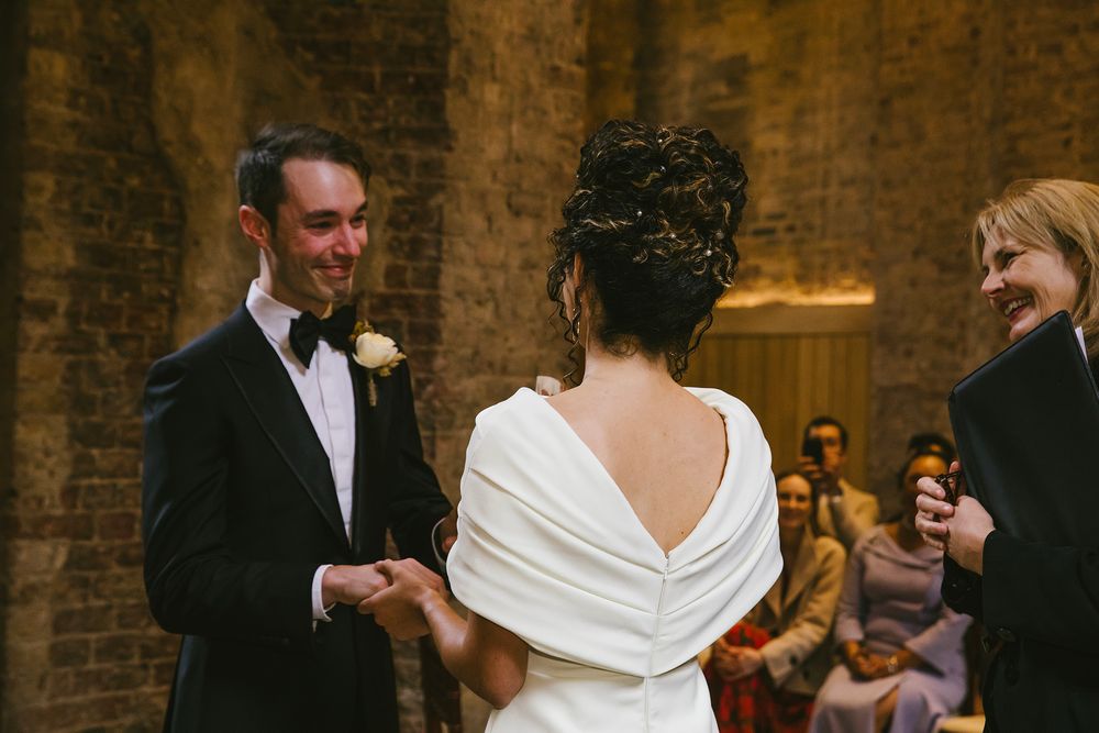 Bride and groom exchange vows at elegant wedding ceremony, bride in white gown with open back, groom in black tuxedo.