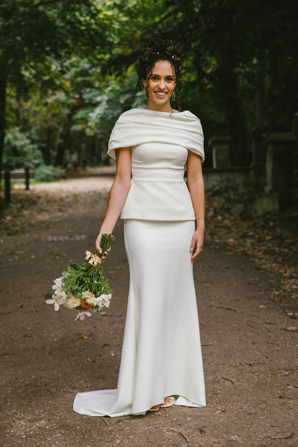 Bride in elegant white cape wedding gown holding floral bouquet, standing on tree-lined path in lush green park.