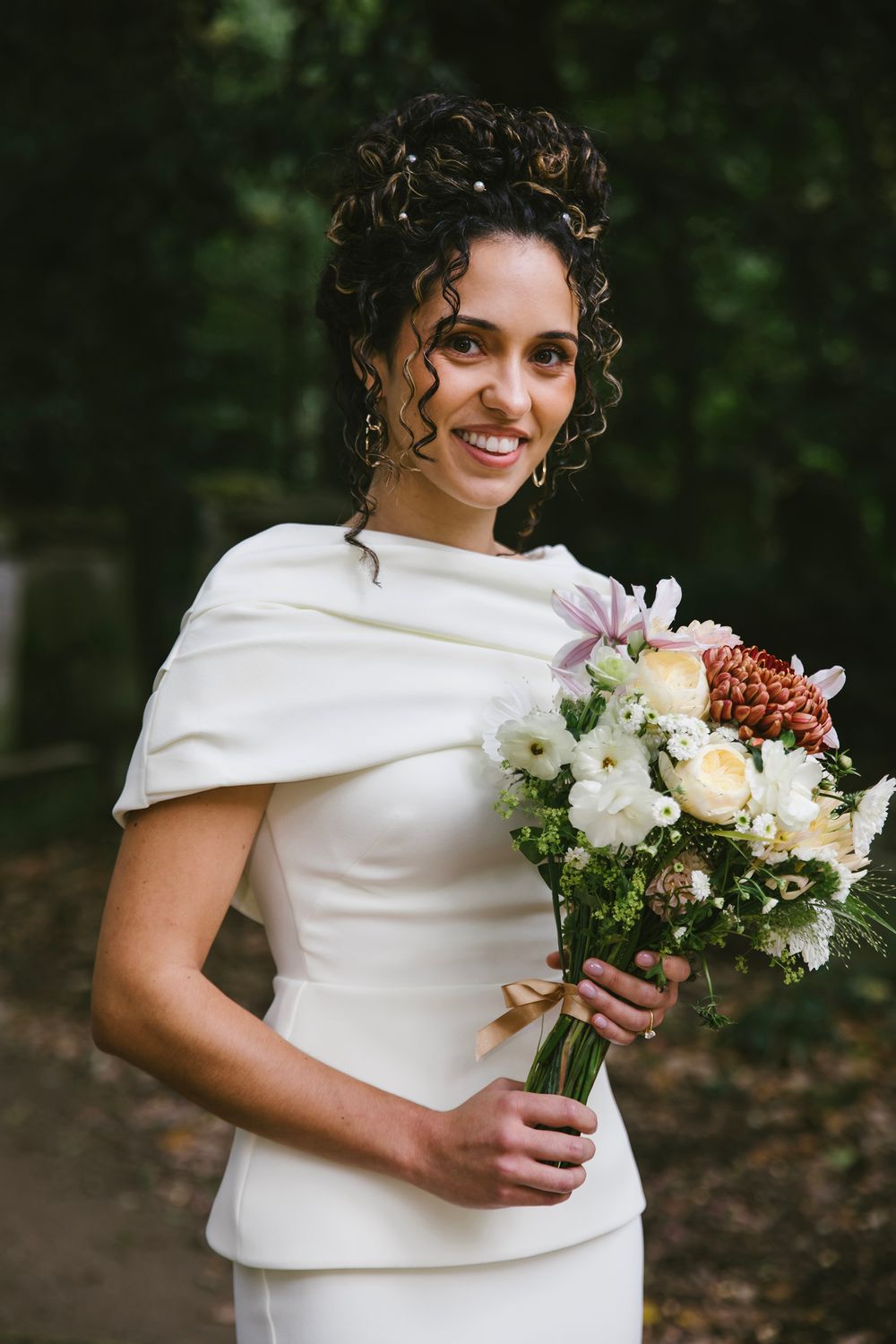 Smiling bride in white cape dress holds a bouquet of roses and wildflowers outdoors in a forest setting.