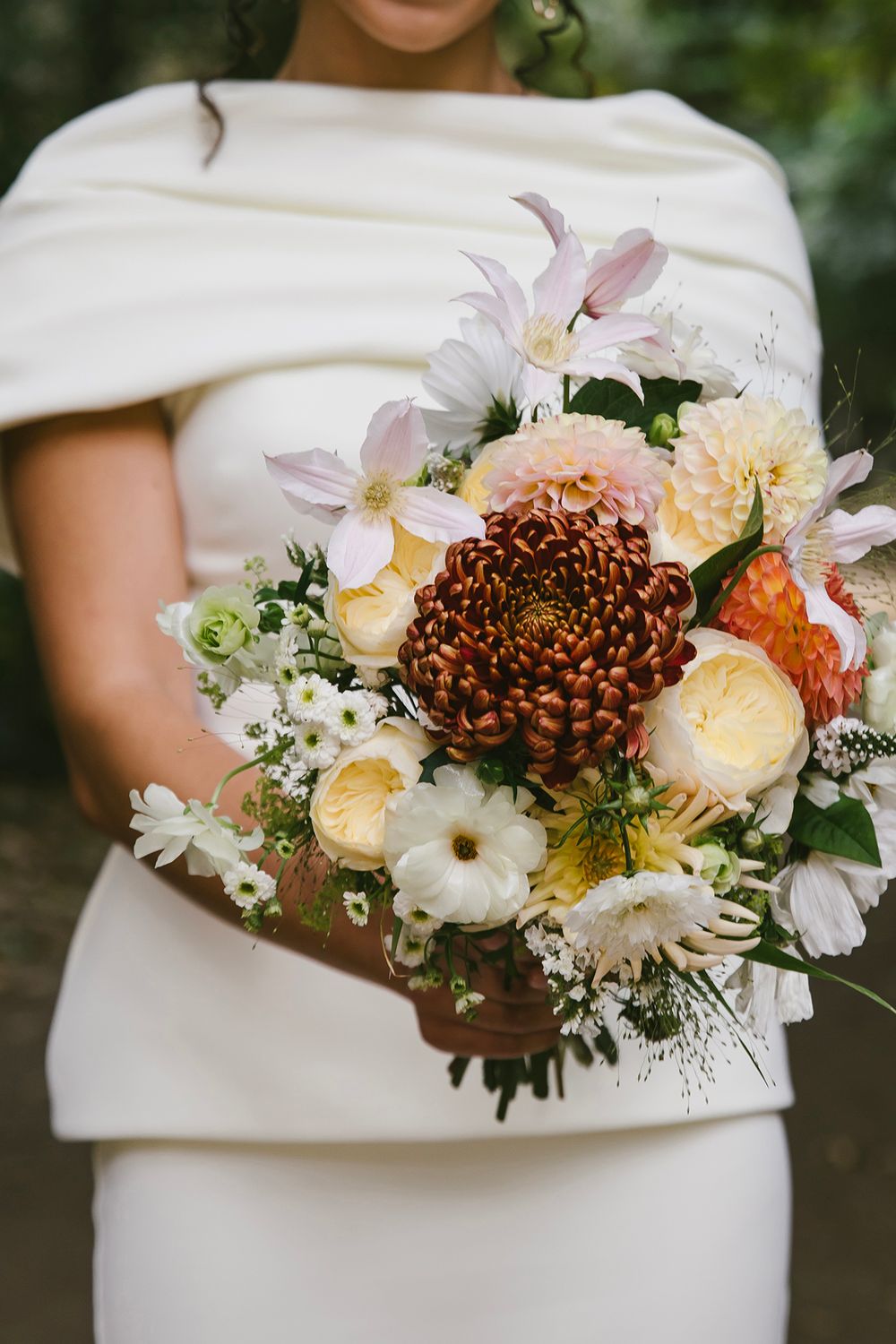Bride in white off-shoulder gown holding lush wedding bouquet with burgundy chrysanthemum, peach dahlias, and white blooms.