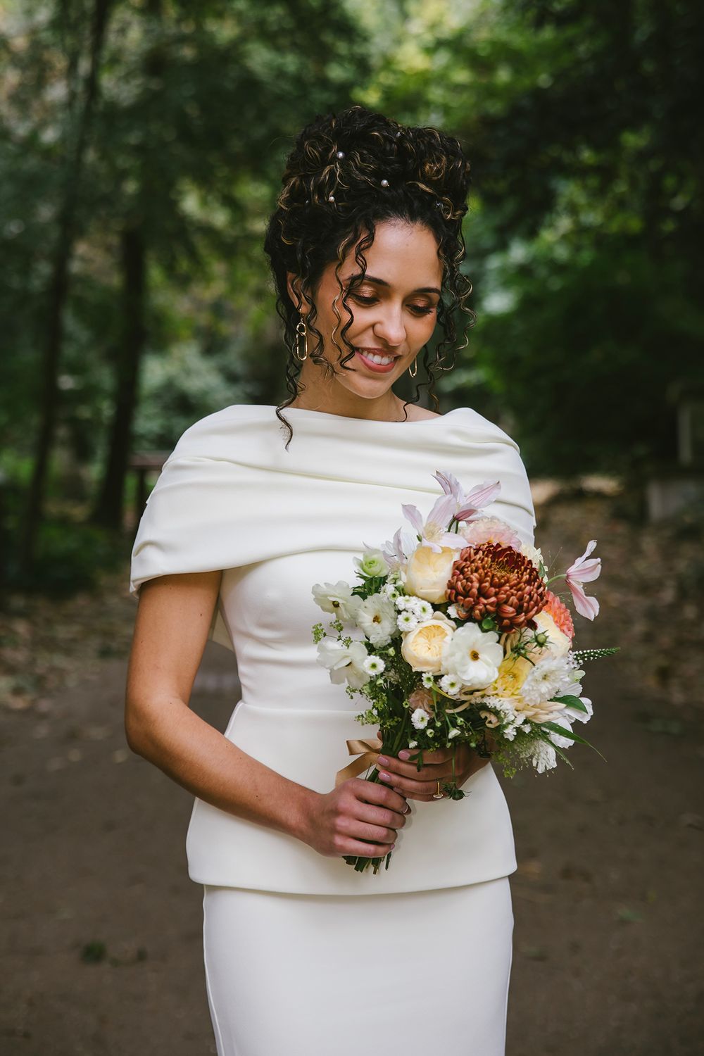 Smiling bride in white cape dress holds colorful bouquet with chrysanthemums outdoors.