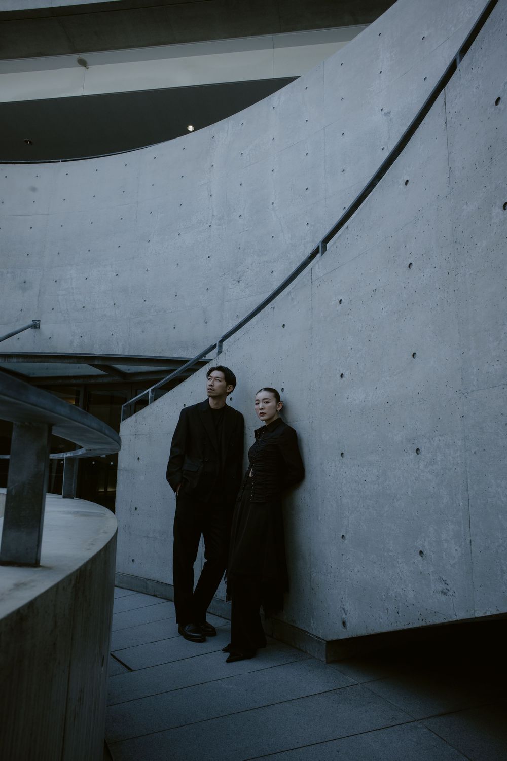 Couple in all-black outfits standing against curved concrete wall of a brutalist architectural space.