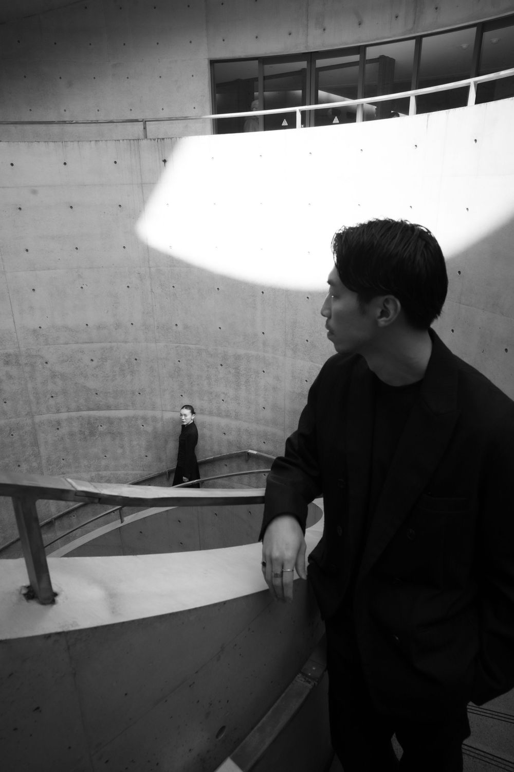 Black and white photo of a man in black jacket leaning on curved concrete railing watching woman below.