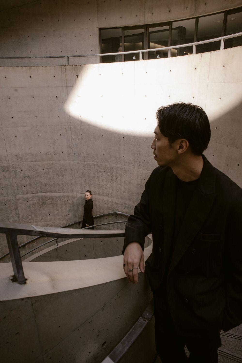 Man in black jacket stands on upper level of spiral concrete structure watching woman on lower level.