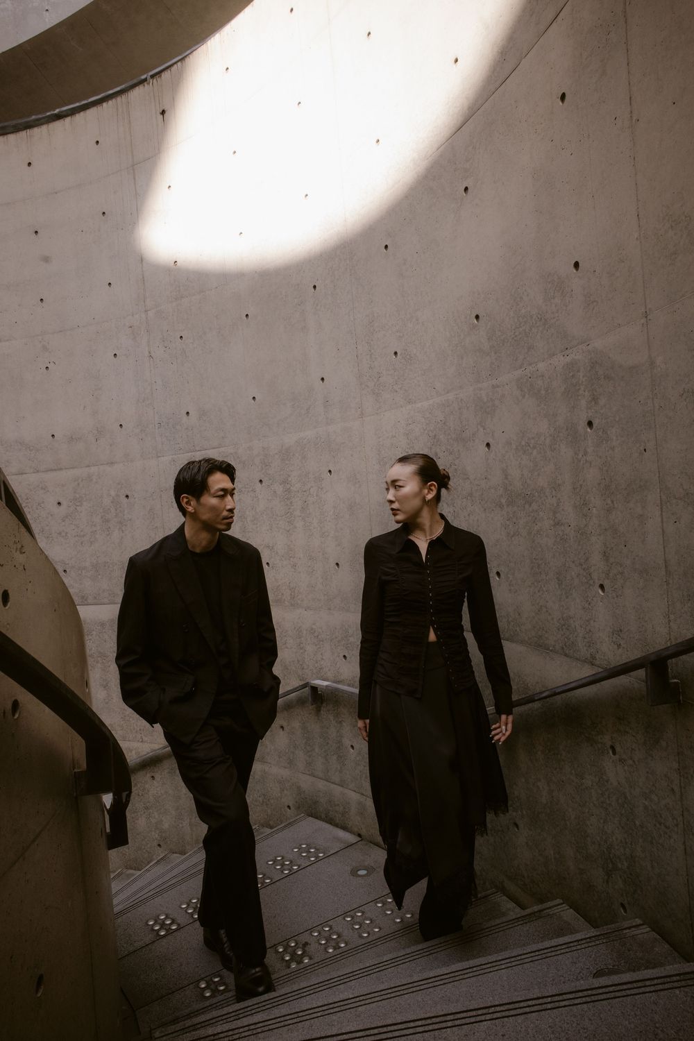 Couple dressed in black walking down spiral concrete staircase in a dramatic brutalist building interior.