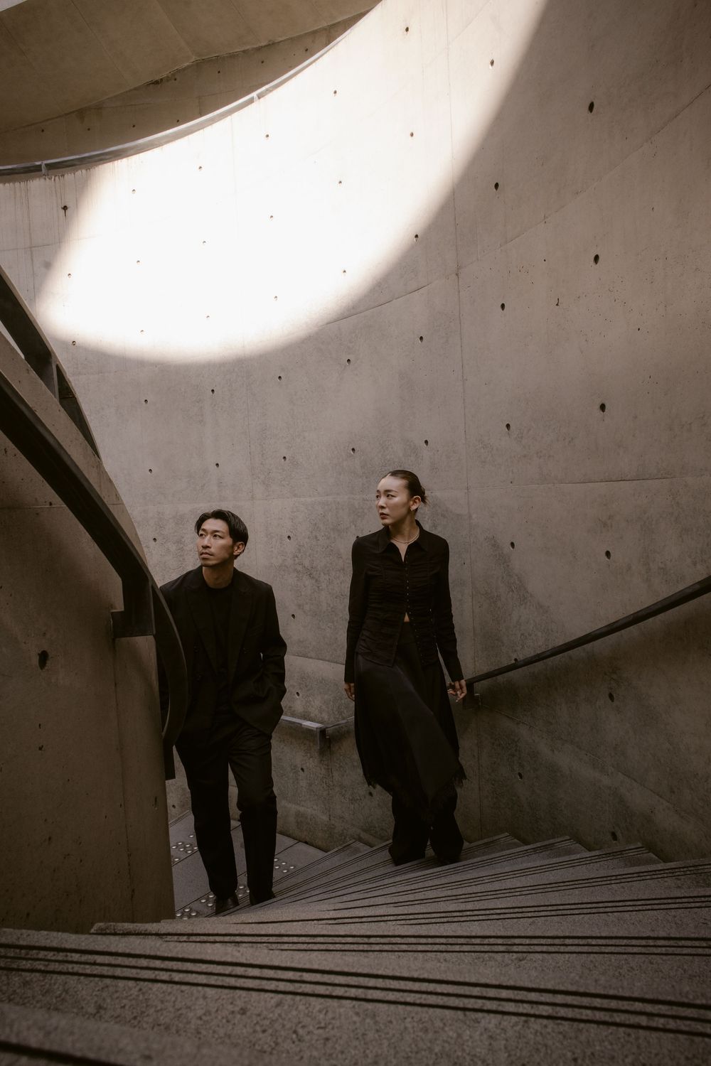 Couple in black outfits descending a curved concrete staircase inside a striking brutalist building.