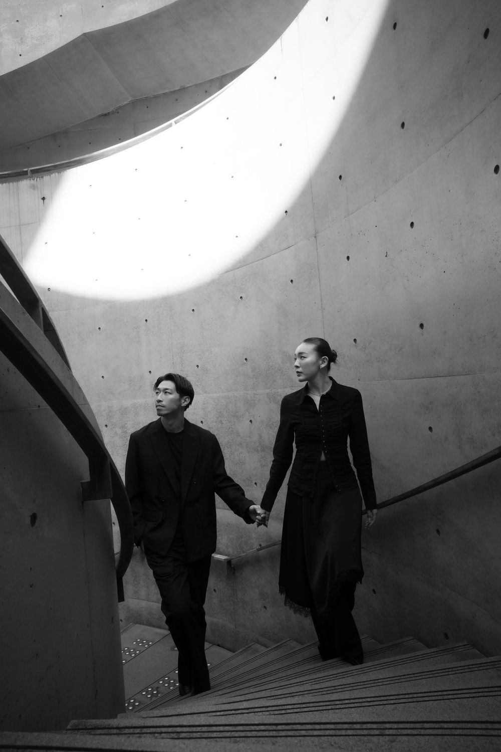 A couple holding hands walks up a dramatic curved concrete staircase in a striking black and white photo.