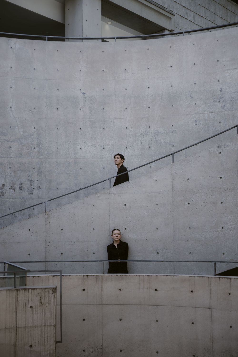 Two people in black coats at different levels of curved concrete staircase against brutalist architecture wall.