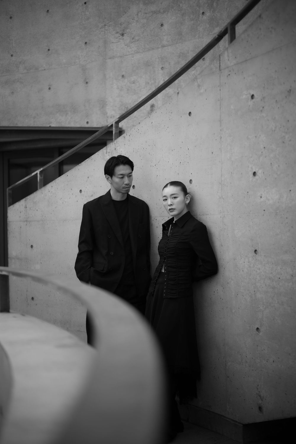 A couple in black outfits pose against a concrete wall near a staircase in a black and white photo.