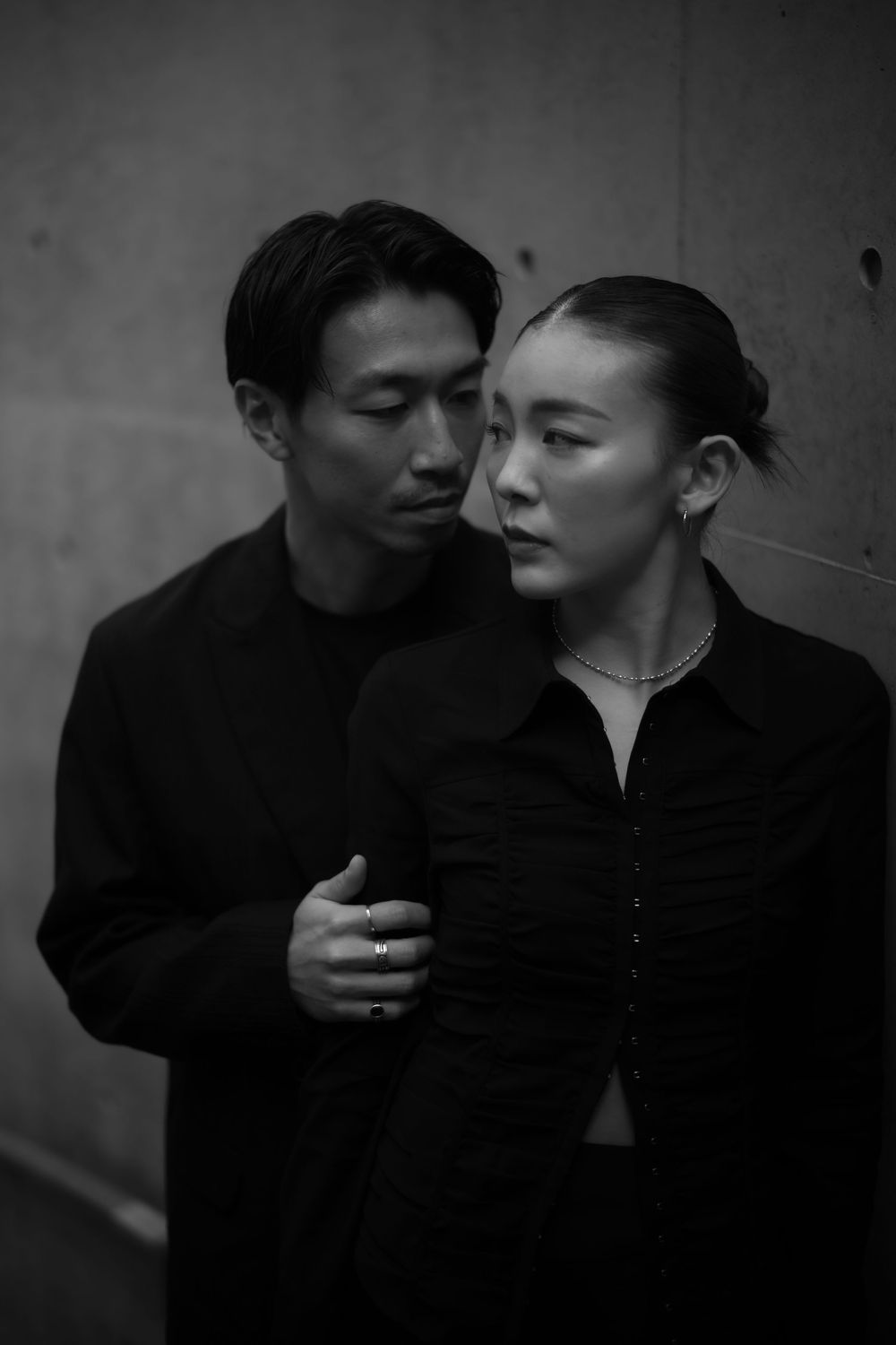 A man embraces a woman from behind against a concrete wall in an intimate black and white portrait.