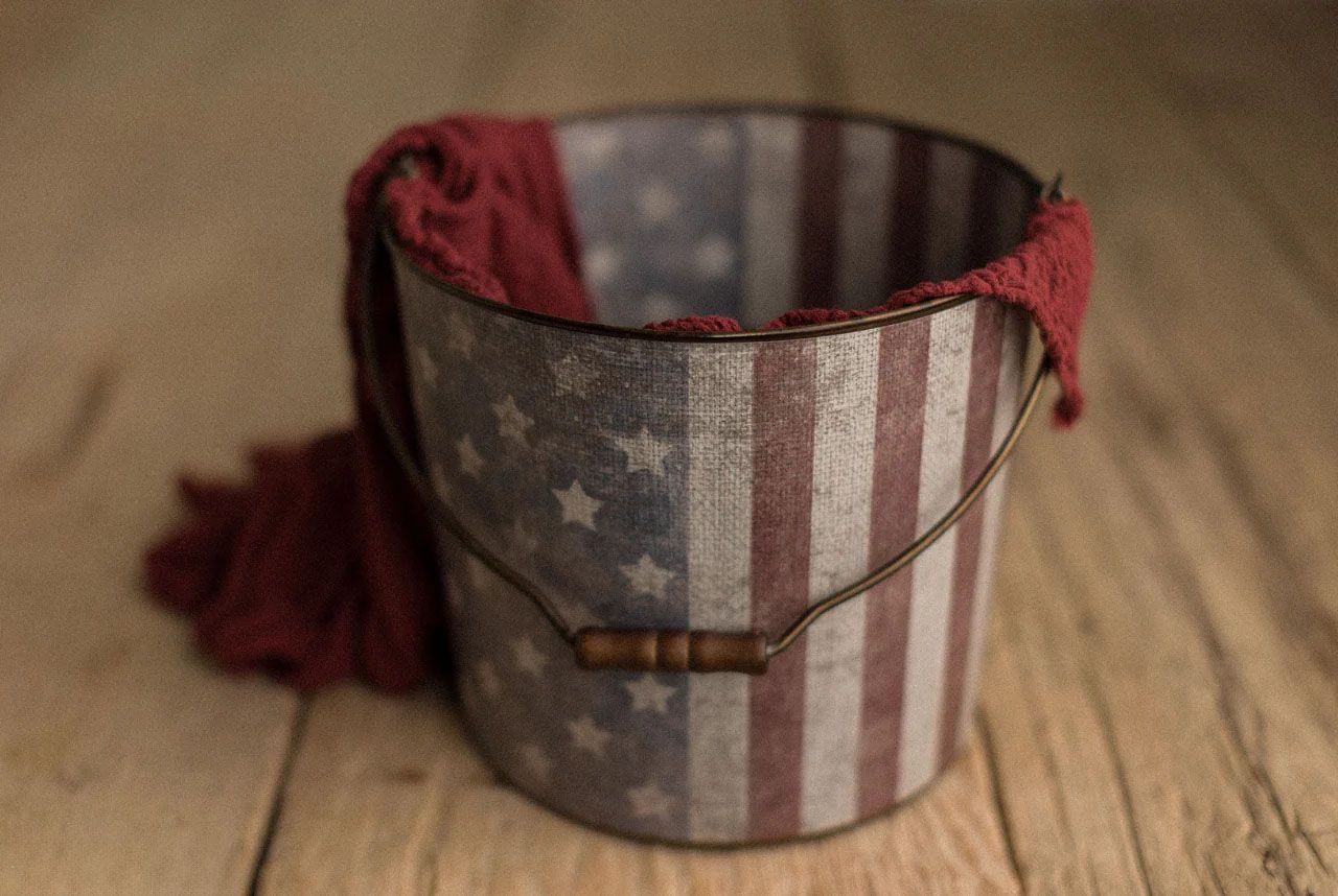 American flag printed metal bucket with red fabric draped inside, sitting on rustic wood surface.