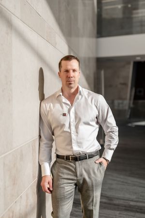 Professional man in white dress shirt and gray trousers leaning against a modern concrete wall indoors.