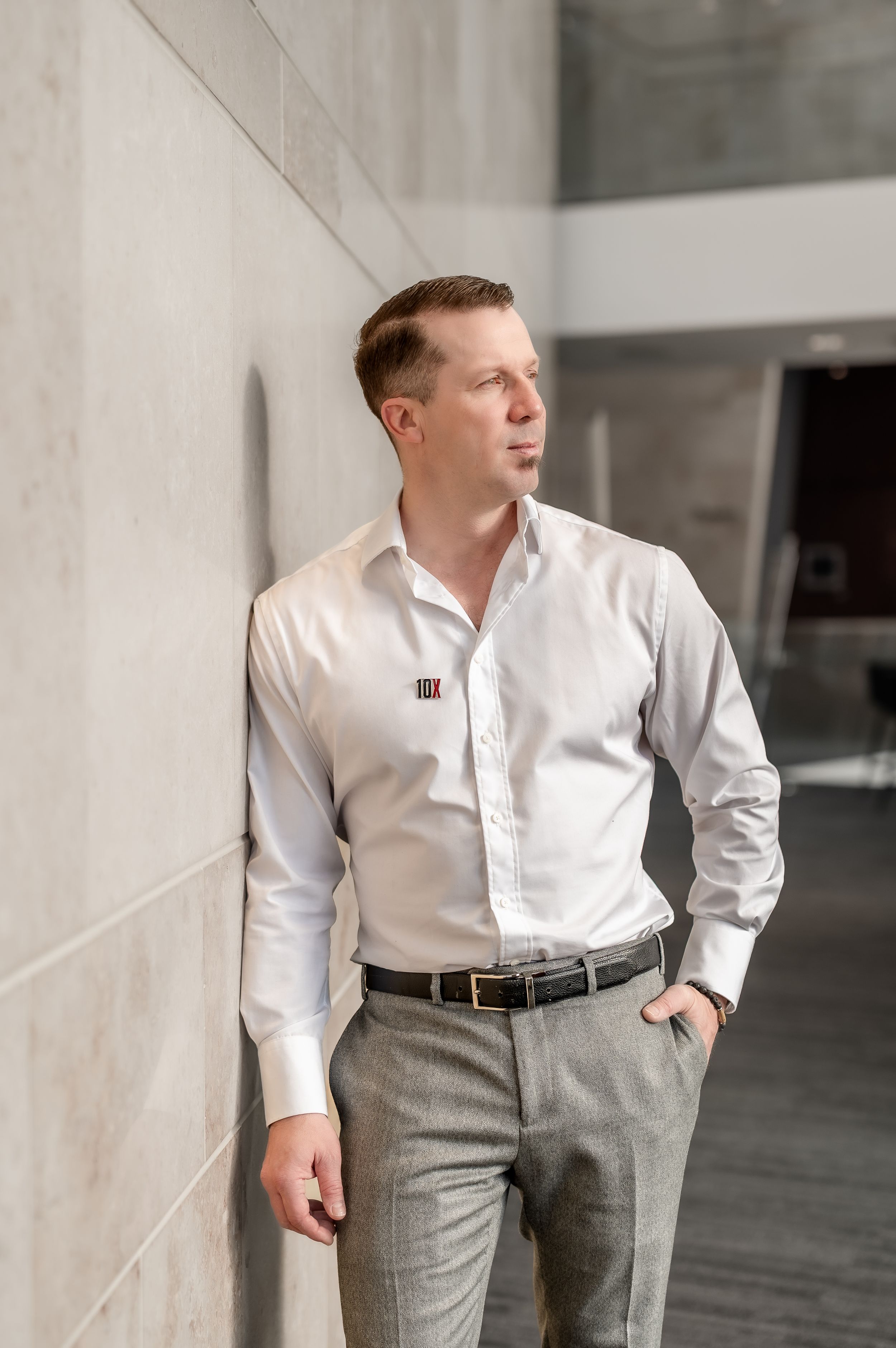 Professional man in white dress shirt and gray trousers leaning against concrete wall in modern office building.