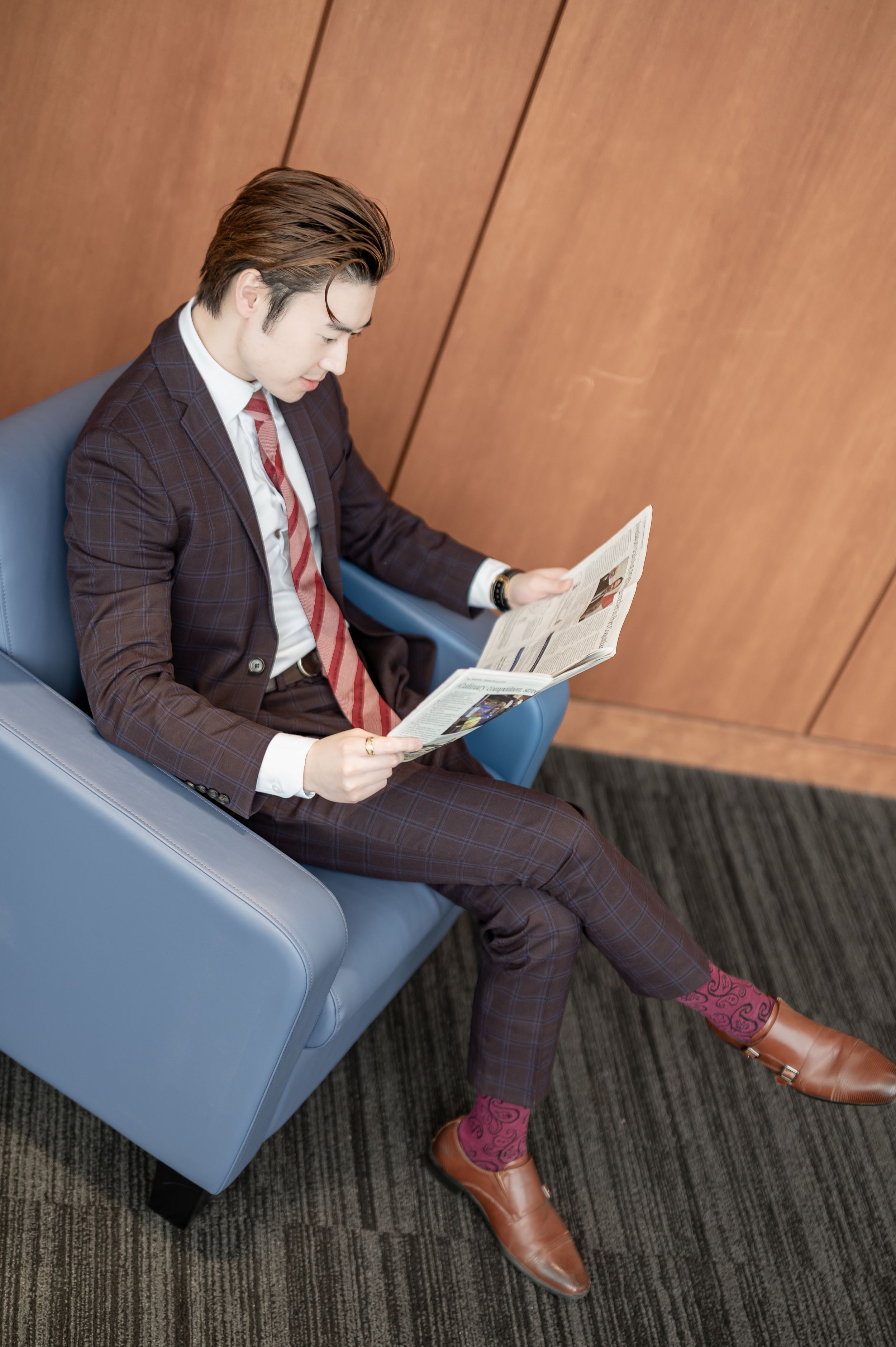 Young businessman in dark suit reading newspaper while seated in blue chair against wooden wall.