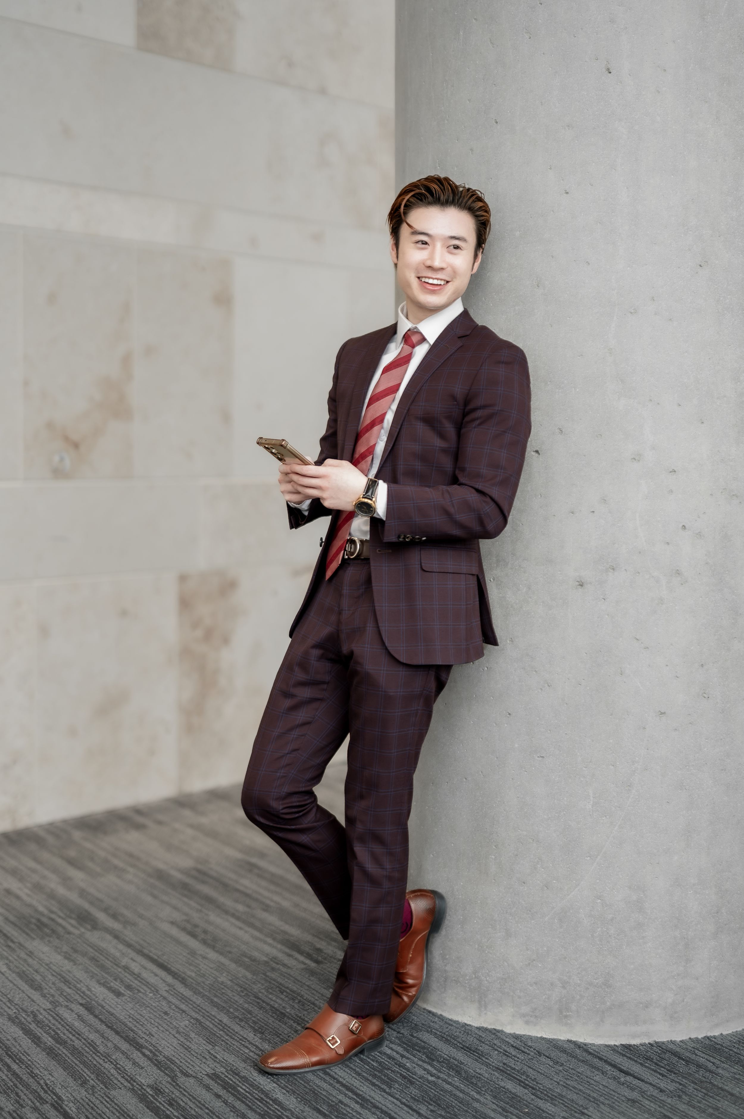 Young Asian businessman in maroon suit holding smartphone, leaning against concrete column, smiling confidently.