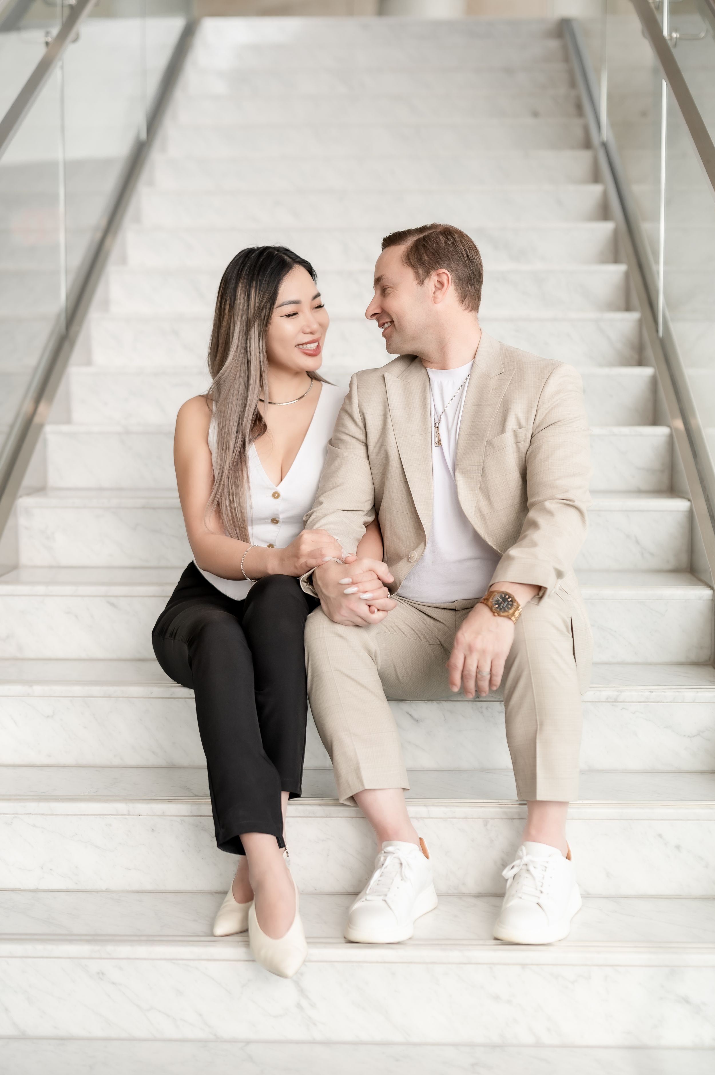 Smiling couple sitting on marble stairs, woman in black pants and man in beige suit holding hands and looking at each other.