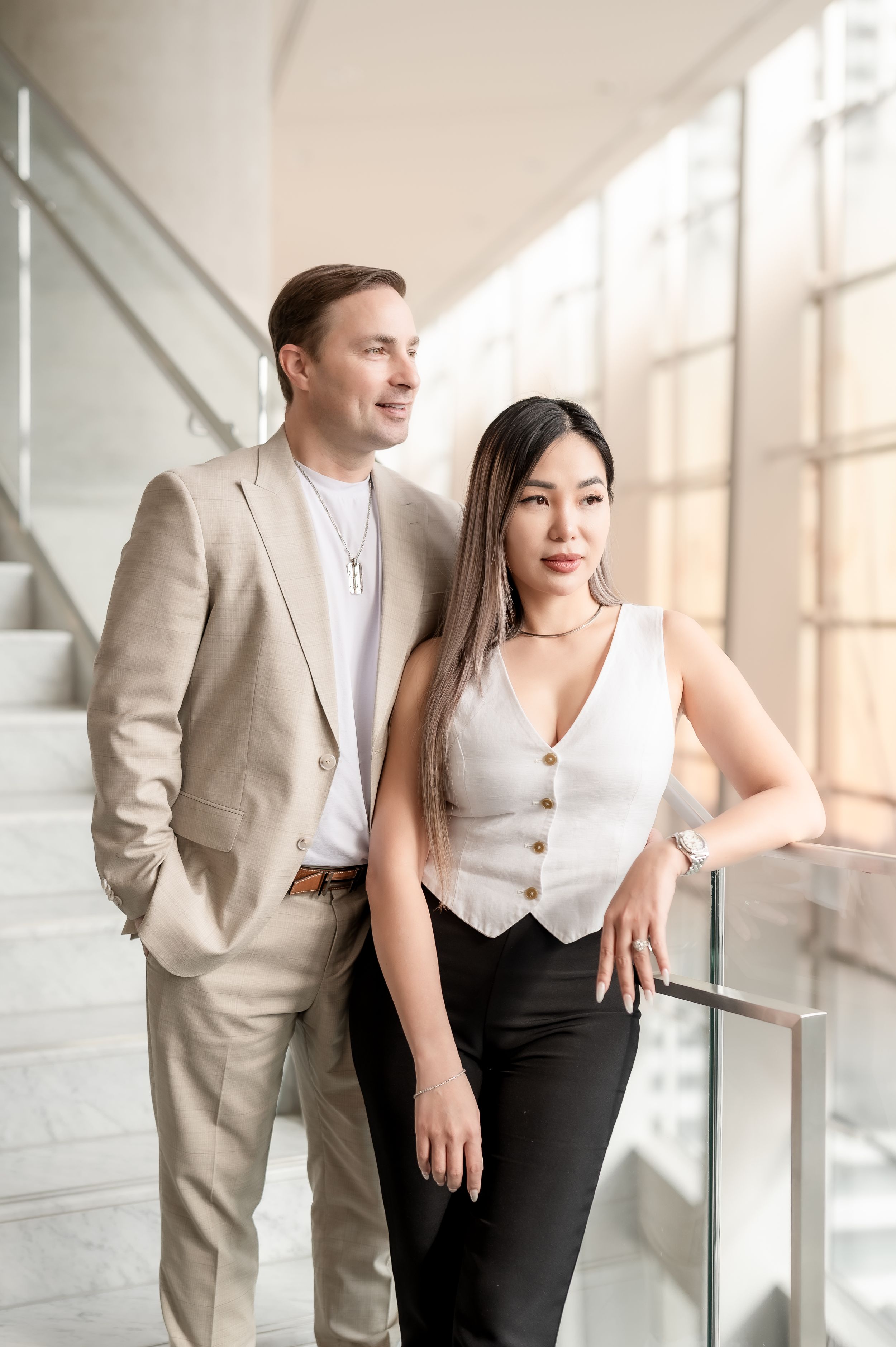 A man in a beige suit and a woman in a white vest and black pants pose elegantly near a modern staircase.