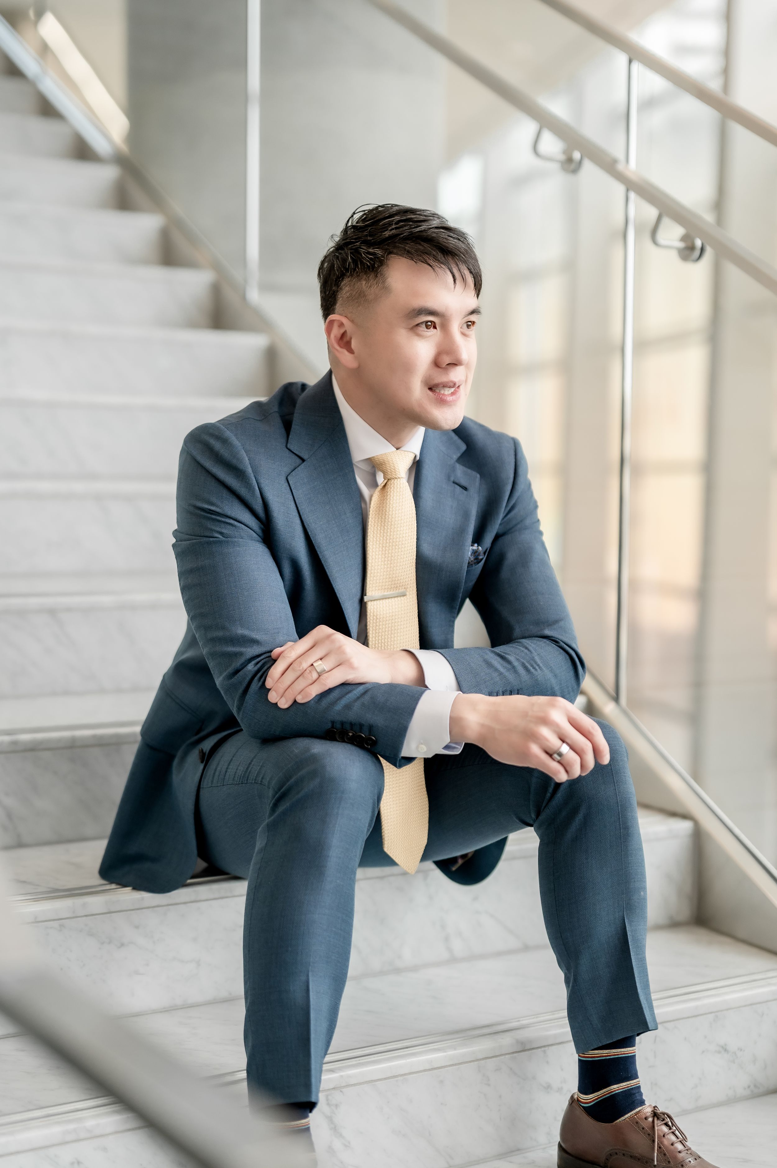Young Asian businessman in blue suit and gold tie sitting on marble stairs in modern building.
