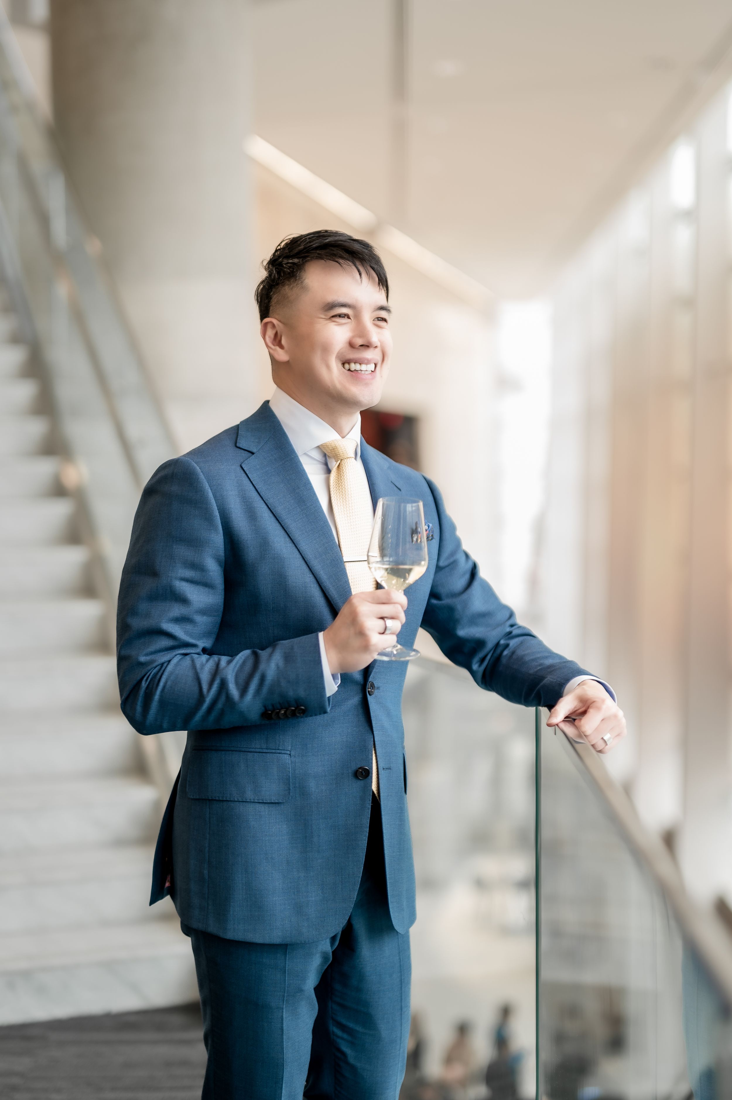Smiling Asian man in blue suit holding white wine glass standing by staircase railing in modern building.