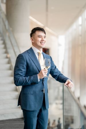 Smiling Asian man in blue suit holding white wine glass standing by staircase railing in modern building.