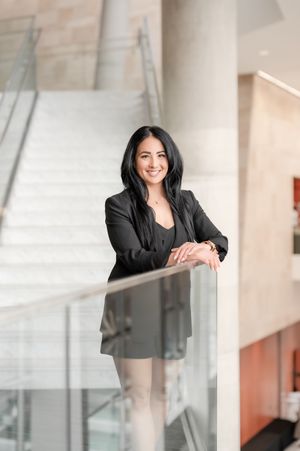 Professional woman in black blazer smiling and leaning on glass railing in modern office building.