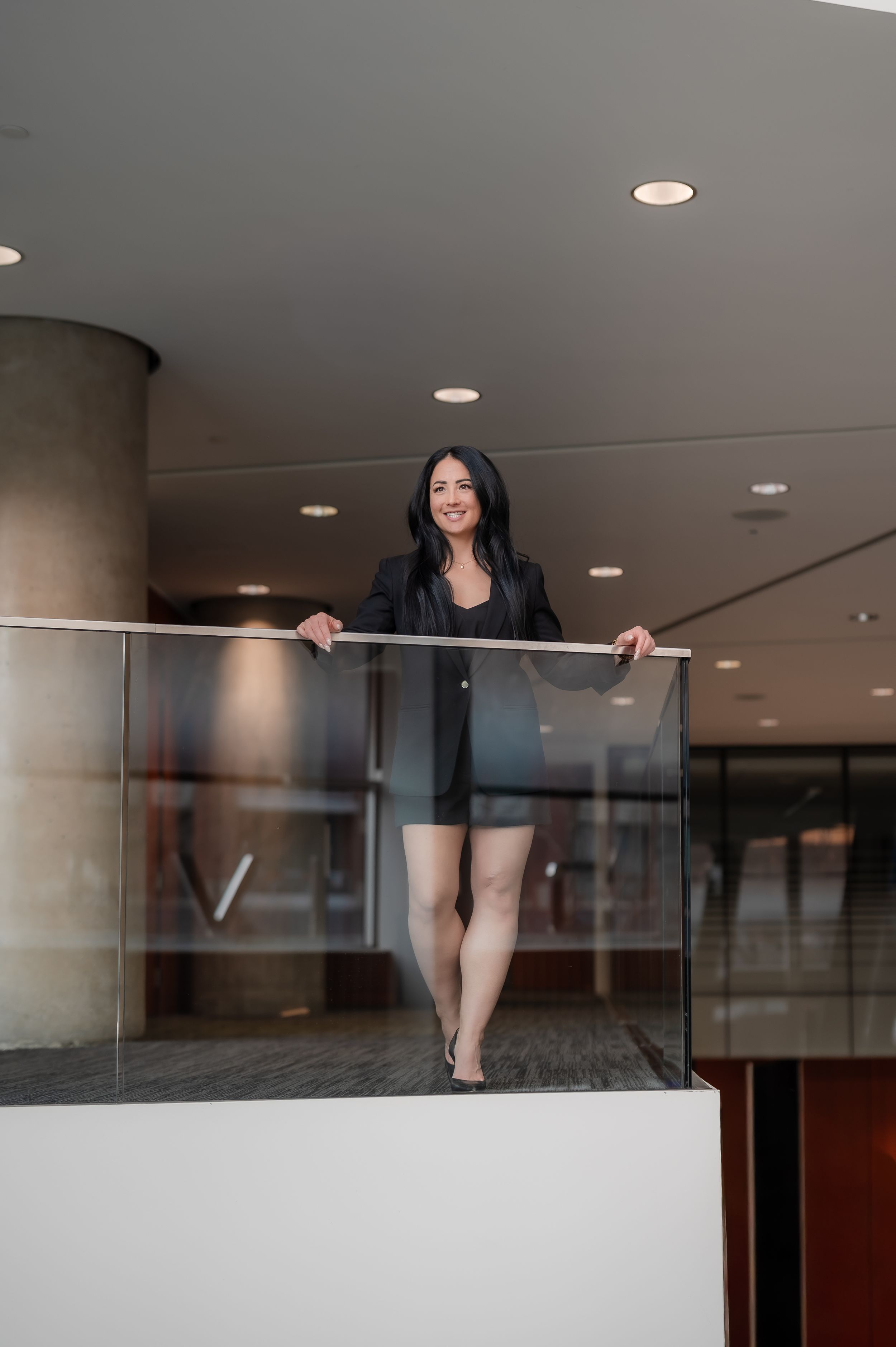 A smiling woman in a black outfit stands confidently at a glass railing on an indoor balcony.
