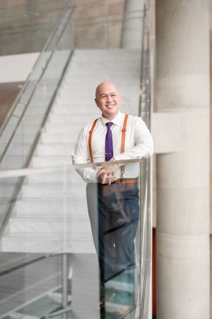 Smiling bald businessman in white shirt, purple tie, and brown suspenders leaning on glass railing in modern building.