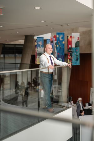 A smiling man in suspenders and jeans leans on a glass railing in a modern building atrium with colorful banners.