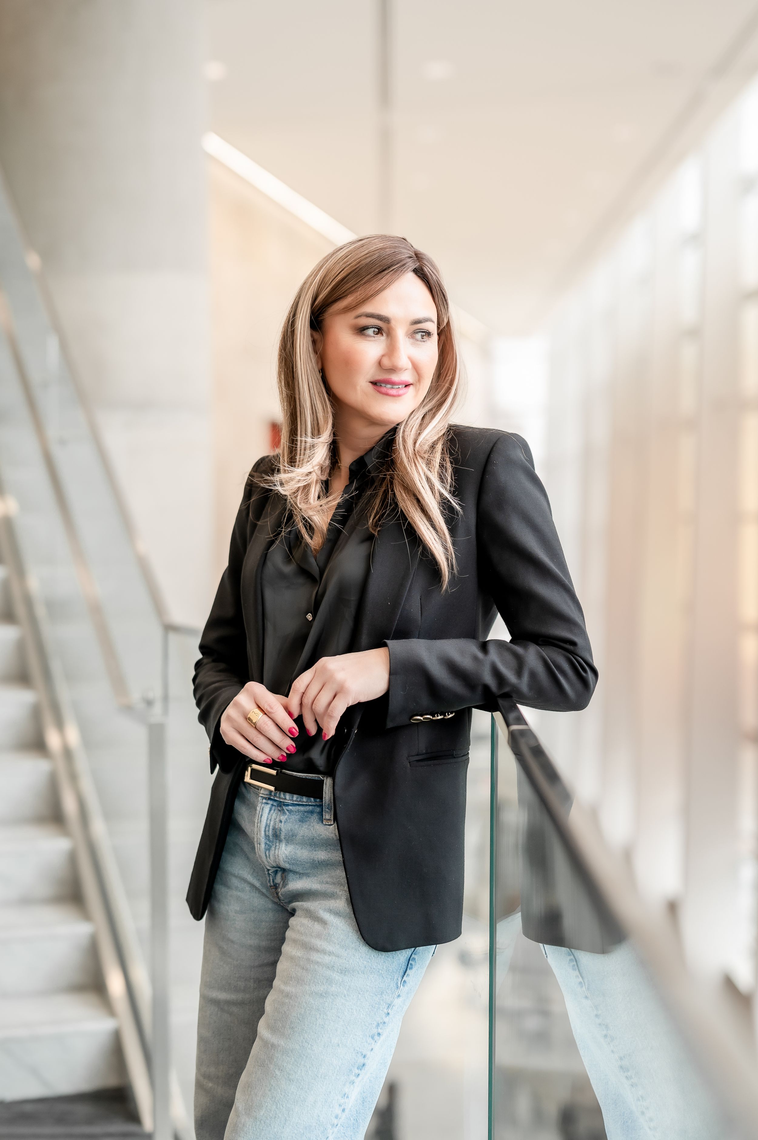 Professional woman in black blazer and jeans leaning on glass railing in modern office building.
