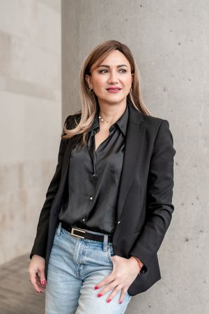 Woman in black blazer and jeans poses confidently against concrete wall, wearing a silk blouse and red nail polish.