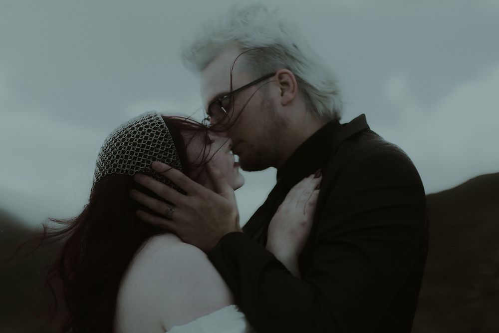 An intimate couple nearly kissing outdoors, woman in lace headpiece and man in black jacket, moody overcast sky.