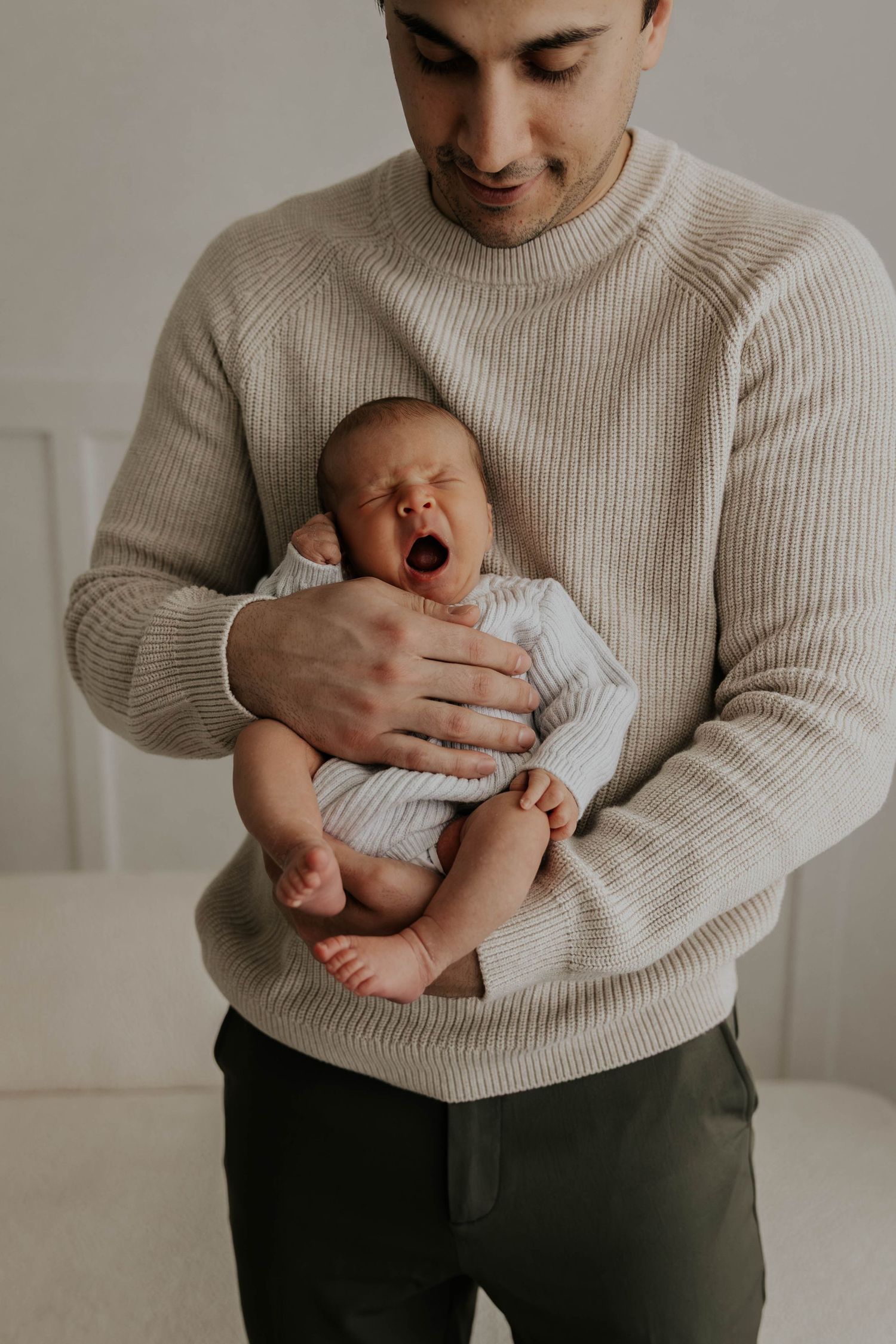 A father in a beige knit sweater tenderly holds his yawning newborn baby against his chest.